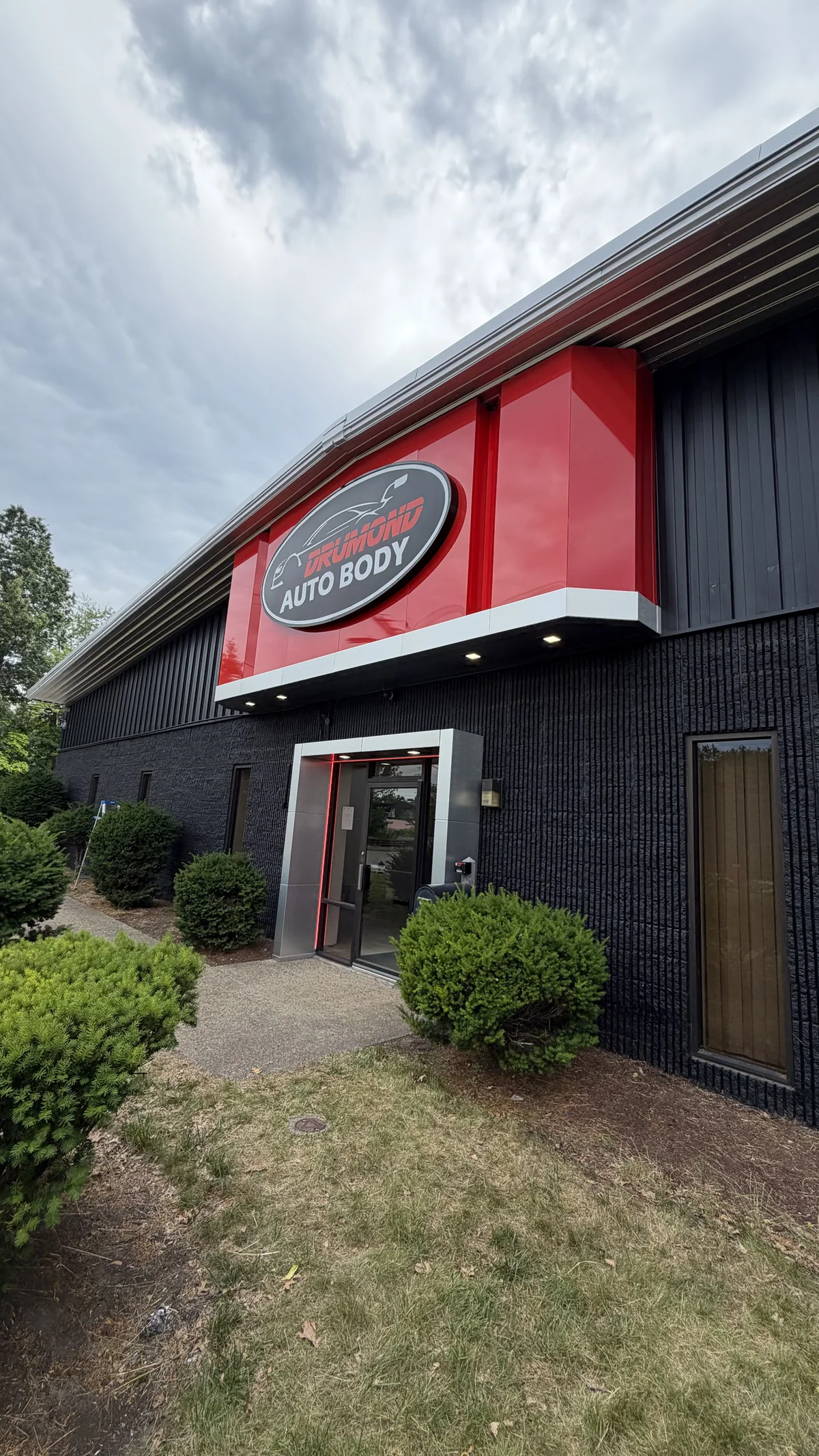 Black building with a red ACM sign box and oval Drumond Auto Body logo above the entrance
